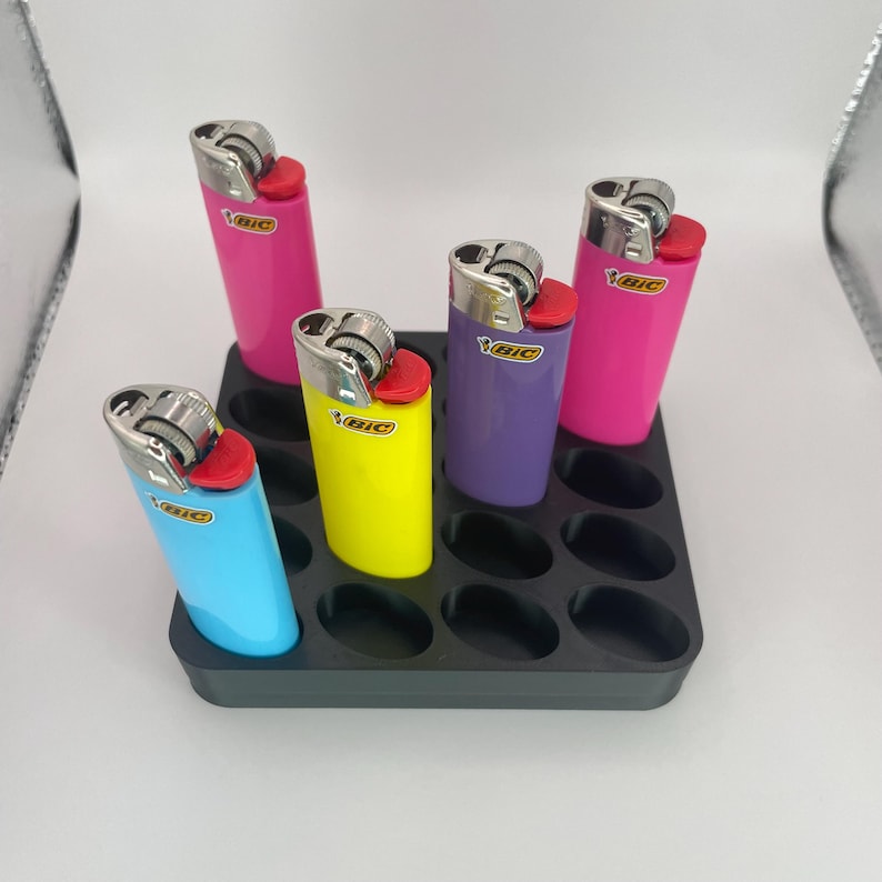 May include: A black plastic organizer holding five colorful BIC lighters. The lighters are in various colors: pink, blue, yellow, and purple. Each lighter has a red ignition button and a silver metal top. The organizer has 12 oval-shaped slots.