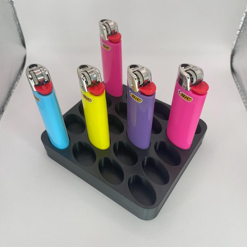 May include: A black plastic lighter holder with six colorful lighters. The lighters are blue, yellow, purple, and pink. The holder has oval-shaped slots to hold the lighters upright.
