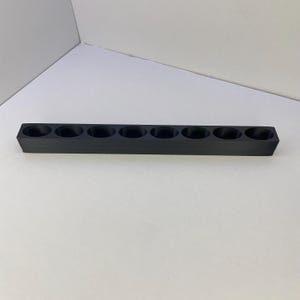 Op de afbeelding: Een lange, rechthoekige zwarte houder met acht gelijkmatig verdeelde ronde openingen in een enkele rij. Het object heeft een effen, matte afwerking en een gestructureerd, lineair ontwerp met een platte basis en rechte zijden.
