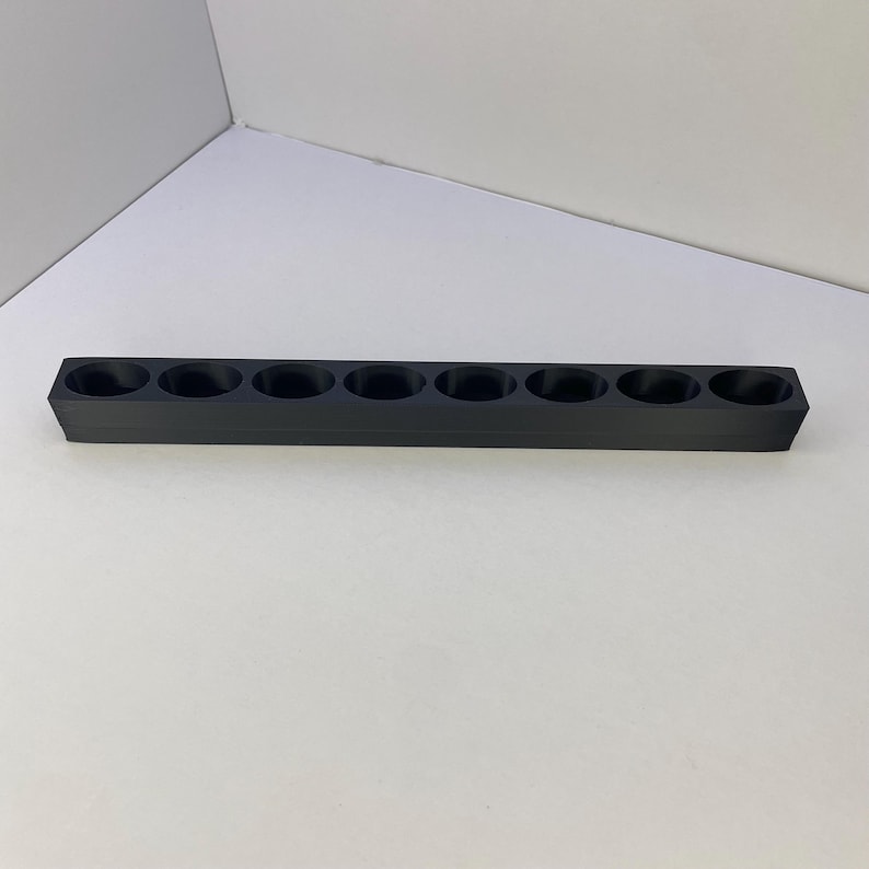 Op de afbeelding: Een lange, rechthoekige zwarte houder met acht gelijkmatig verdeelde ronde openingen in een enkele rij. Het object heeft een effen, matte afwerking en een gestructureerd, lineair ontwerp met een platte basis en rechte zijden.