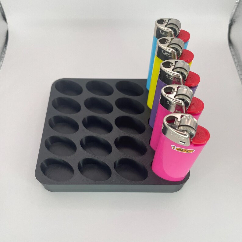 May include: A black plastic lighter holder with oval compartments. Six colorful lighters are in the holder, including blue, yellow, purple, and pink. The lighters are arranged in a row, with the holder on a white surface.