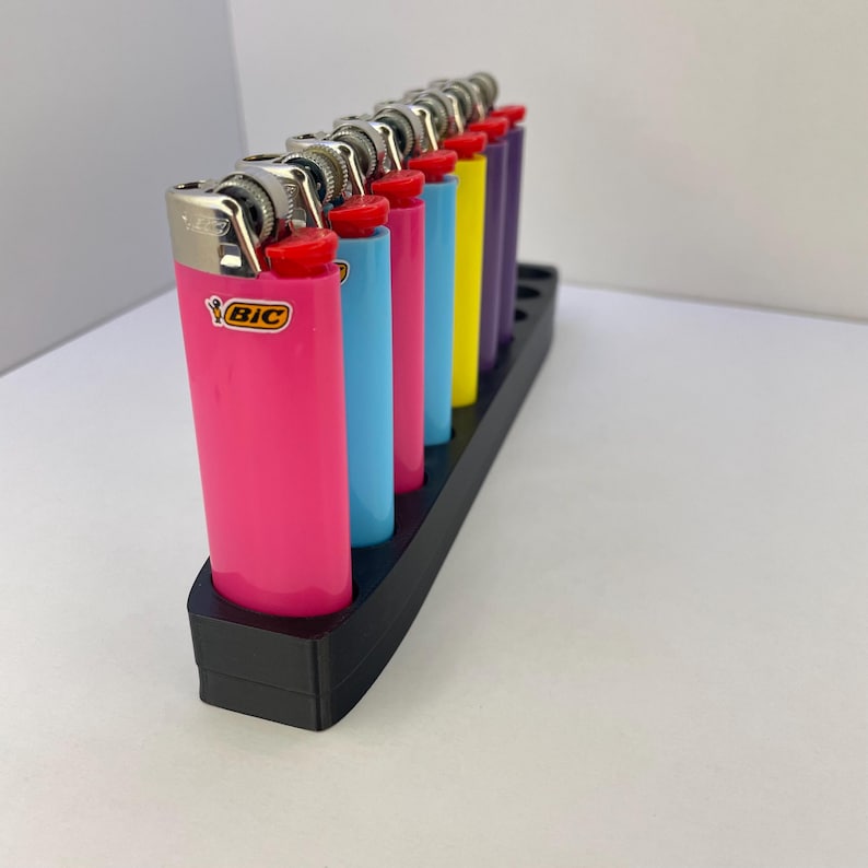 Pu&ograve; includere: Una collezione di sette accendini Bic di vari colori, tra cui rosa, azzurro, magenta, ciano, giallo, viola e blu scuro, disposti in fila all'interno di un supporto rettangolare in plastica nera con fori circolari per ogni singolo accendino.