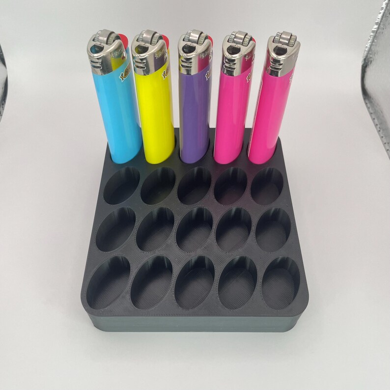 May include: A black plastic lighter holder with 18 oval slots, holding five colorful lighters. The lighters are blue, yellow, purple, and two shades of pink. The lighters have silver metal tops and are arranged in a neat row.