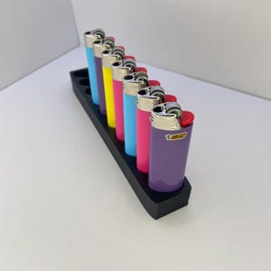 K&ouml;nnte beinhalten: Eine Reihe von sieben bunten Bic-Feuerzeugen in einem schwarzen rechteckigen Halter. Die Feuerzeuge haben metallisch silberne Oberteile und Geh&auml;use in den Farben Hellblau, Violett, Gelb, Pink, Hellblau, Dunkelpink und Violett mit dem Bic-Logo.