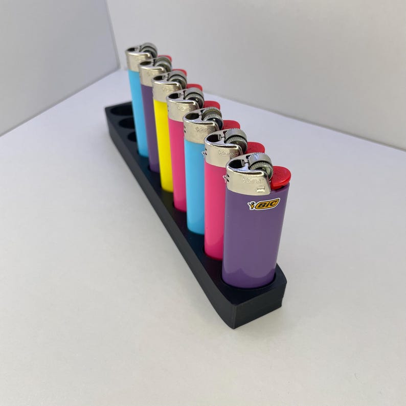 K&ouml;nnte beinhalten: Eine Reihe von sieben bunten Bic-Feuerzeugen in einem schwarzen rechteckigen Halter. Die Feuerzeuge haben metallisch silberne Oberteile und Geh&auml;use in den Farben Hellblau, Violett, Gelb, Pink, Hellblau, Dunkelpink und Violett mit dem Bic-Logo.