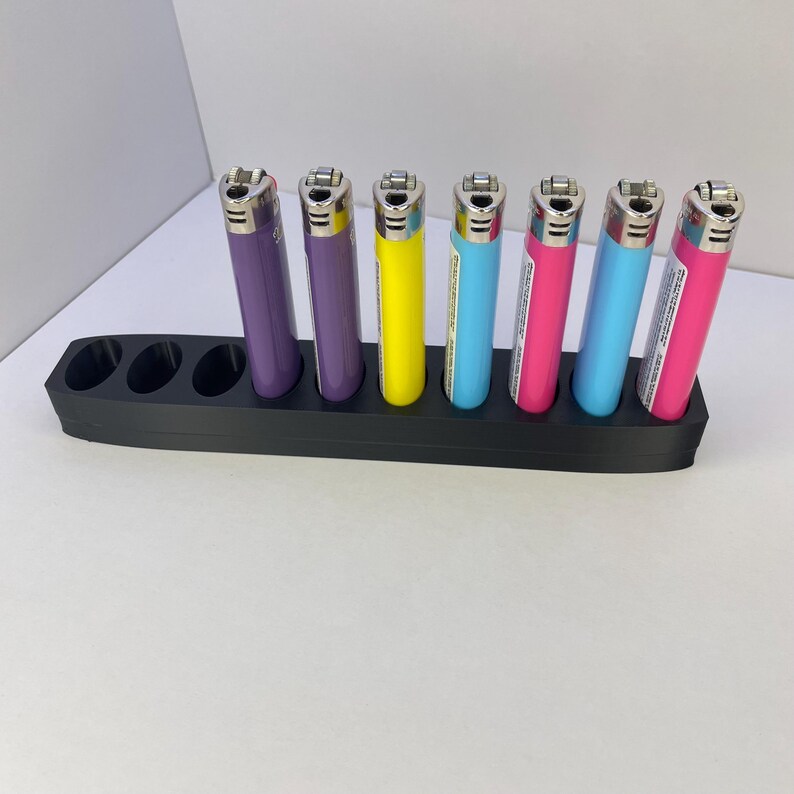 Pu&ograve; includere: Un supporto rettangolare in plastica nera con sette fori circolari, contenente sei accendini colorati viola, gialli, azzurri e rosa. Il supporto presenta tre fori vuoti sul lato sinistro, mentre gli accendini sono disposti in verticale negli spazi rimanenti.