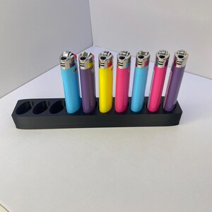 K&ouml;nnte beinhalten: Ein schwarzer, rechteckiger Organizer-St&auml;nder mit sieben runden Aussparungen, in dem sieben bunte Feuerzeuge in Hellblau, Lila, Gelb, Pink und Blau stecken. Jedes Feuerzeug hat einen silbernen Z&uuml;ndkopf und einen schlanken, zylindrischen K&ouml;rper.