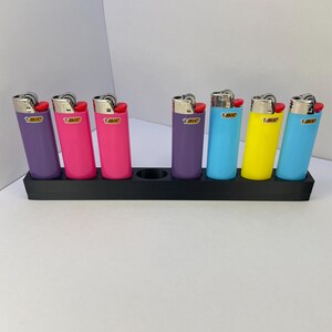 Op de afbeelding: Een zwarte rechthoekige standaard met zeven BIC-aanstekers in verschillende kleuren, waaronder paars, roze, blauw en geel. Elke aansteker heeft een metalen bovenkant, een rode ontstekingsknop en het BIC-logo op de plastic behuizing.
