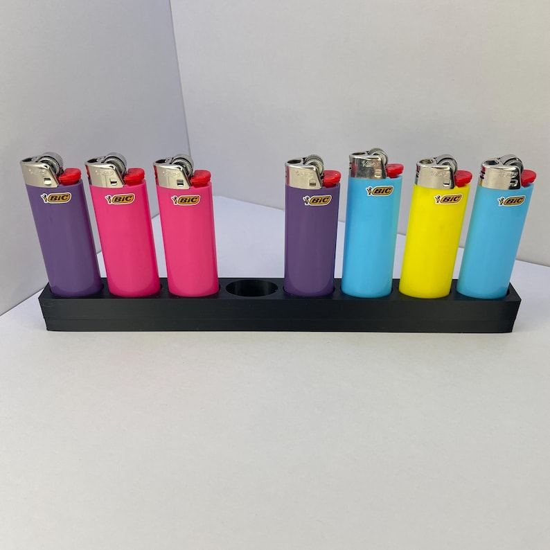 Op de afbeelding: Een zwarte rechthoekige standaard met zeven BIC-aanstekers in verschillende kleuren, waaronder paars, roze, blauw en geel. Elke aansteker heeft een metalen bovenkant, een rode ontstekingsknop en het BIC-logo op de plastic behuizing.