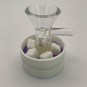 May include: A clear glass bowl with a handle, set inside a white, circular container with a purple interior. The bowl holds several white, cube-shaped objects. The container is on a white surface.