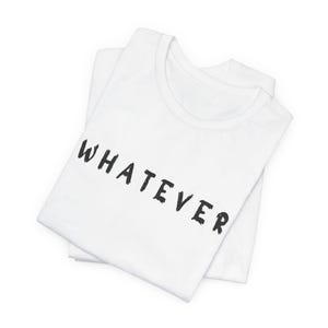 May include: Two white t-shirts are folded and stacked. The top shirt has the word "WHATEVER" printed in black, brush-style lettering across the front. The shirts are on a white background.