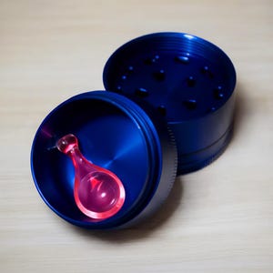 May include: A blue metal grinder with a pink scoop inside. The grinder is open, revealing the interior and the scoop. The grinder has a circular shape with a textured exterior. The scoop is pink and has a rounded shape.