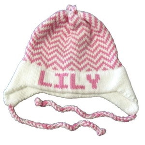 May include: White and pink knitted baby hat with a chevron pattern and the name "LILY" embroidered on the front. The hat has a long, pink and white braided tie.