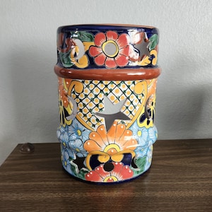 May include: A hand-painted ceramic candle holder with a vibrant floral design. The cylindrical holder features a blue, orange, and red colour scheme with cut-out star and flower shapes. The top and bottom rims are brown.