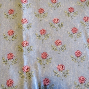 May include: A light blue fabric with a repeating pattern of pink roses and green leaves. The roses are in full bloom and are evenly spaced across the fabric. The background has a subtle, textured appearance.