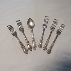 May include: A set of five silver-toned metal utensils consisting of four forks and one spoon. Each piece features an ornate, embossed floral pattern along the handle and a polished finish. The items are arranged in a fan-like formation.