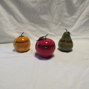 May include: Three ceramic fruit-shaped containers with lids. One is a yellow-orange textured orange, one is a glossy red apple, and one is a green pear. Each piece features a dark metal stem and leaf detail attached to the top of the lid.
