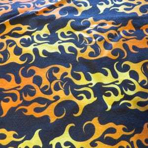 May include: Close-up of a fabric with a repeating flame pattern. The flames are in shades of orange and yellow against a dark blue background. The fabric has a slightly textured appearance.
