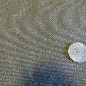 May include: Close-up of a textured fabric in shades of olive green and tan. A U.S. quarter coin is placed on the fabric for scale. The fabric has a woven appearance with a subtle pattern.