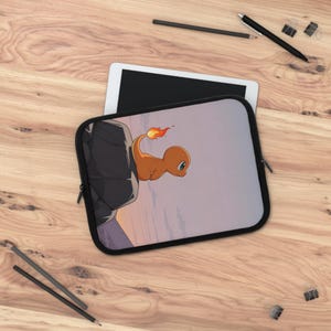 Puede incluir: Funda negra para portátil con un personaje Charmander naranja con cola de llama, sobre un fondo de cielo púrpura y rosa. La funda tiene una cremallera y un borde negros.