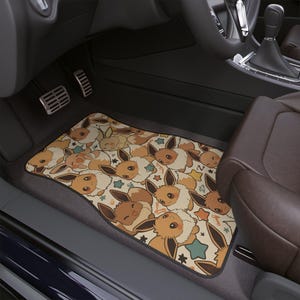 May include: A rectangular car floor mat featuring a repeating pattern of brown and cream-colored Eevee characters with large eyes and pointed ears, interspersed with small teal and yellow stars on a light beige background with a black border.