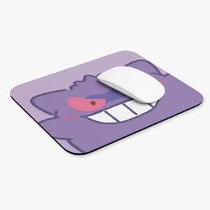 May include: A purple mousepad featuring a cartoon image of a Gengar character with a white mouse on top. The mousepad has a black border, and the Gengar has red eyes and a wide grin.