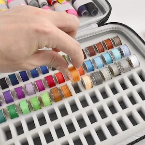 May include: A portable storage case containing numerous small, clear plastic bobbins wound with sewing thread in a wide variety of colors. The bobbins are organized in a foam insert with individual rectangular slots for each spool.