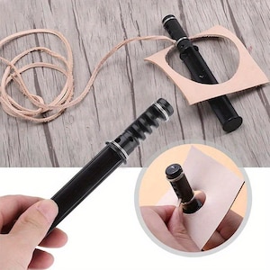 Peut inclure: Outils de maroquinerie noirs avec des détails argentés, dont un cutter rotatif et une lanière en cuir. Un morceau de cuir beige est découpé en forme carrée. Les outils sont sur une surface en bois.
