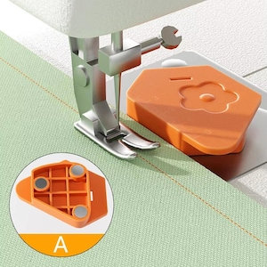 May include: A sewing machine with a silver presser foot and an orange magnetic seam guide. The seam guide has a flower design and is positioned near the needle. The fabric is light green with a stitched seam.