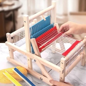 Könnte beinhalten: Ein Holzwebstuhl mit einem roten und blauen Schussfaden und einem weißen Kettfaden. Der Webstuhl ist zum Weben eingerichtet und es liegen Knäuel mit gelbem und blauem Garn auf dem Tisch.