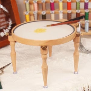 May include: A wooden embroidery stand with a round frame and three legs. The stand holds a white fabric with yellow embroidery. A needle and thread are placed on the fabric. The stand is surrounded by spools of thread.