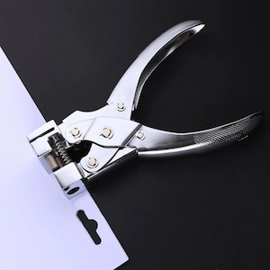 May include: A silver-colored metal hole punch tool with a spring-loaded mechanism and textured handles. The tool is positioned over a white cardstock sheet that features a pre-cut elongated slot, showing the functional design of the handheld device.