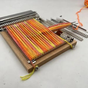 May include: A wooden hand loom with a metal comb and tensioning bar, featuring vertical warp threads in a gradient of yellow, orange, and pink. The loom includes a metal shuttle and additional metal components, with loose orange yarn extending from the side.