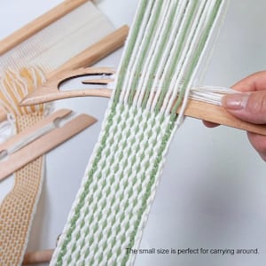 Puede incluir: Una lanzadera de madera se utiliza para tejer una tira de tela con hilo verde salvia y blanco alternado. En el fondo, se observa un marco de telar de madera y una tira parcialmente tejida de hilo amarillo y blanco. El texto dice: The small size is perfect for carrying around.