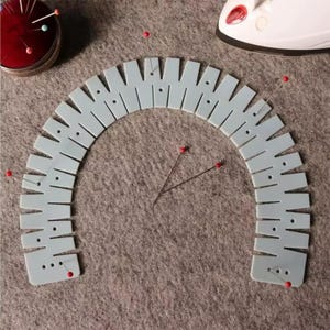 Peut inclure: Un gabarit de couture en plastique bleu clair en forme de demi-cercle avec des fentes et des petits trous régulièrement espacés, accompagné de plusieurs épingles à tête rouge et bleue et d'un pelote à épingles rouge.
