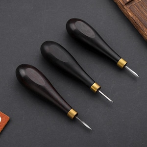 Può includere: Tre lesine per la lavorazione della pelle con manici in legno scuro e puntali in ottone. Le lesine hanno una lama appuntita color argento. Gli strumenti sono disposti diagonalmente su una superficie grigio scuro, suggerendo la lavorazione della pelle o l'artigianato.