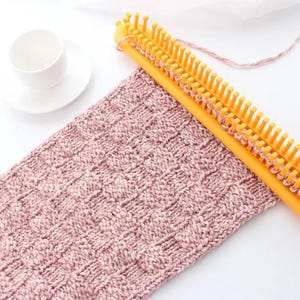 May include: A yellow plastic knitting loom with a row of pegs holds a piece of textured, dusty pink yarn fabric. The knitted fabric features a repeating square pattern. A small white ceramic cup and saucer are positioned to the side of the loom.