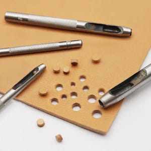 May include: A close-up of leather crafting tools. Several silver metal hole punch tools are arranged on a tan leather surface. The leather has multiple punched holes and small circular leather cutouts. The tools are used for leather working.