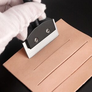 May include: A leather craft skiving tool with a wide, flat, silver-toned metal blade and a black handle mount secured by two silver screws. The tool is positioned on a square piece of light tan leather that features three parallel straight-line cuts.