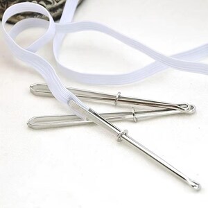 May include: Three silver-toned metal cake lifters with a white elastic band. The lifters have a long, slender handle and a circular ring near the top. The elastic band is white and appears to be for securing the lifters.
