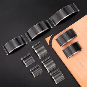 May include: A collection of leathercraft tools, including various sizes of curved and rectangular steel cutters. A small piece of tan leather is also present, suggesting the tools' use in leatherworking projects. The tools are arranged on a black surface.