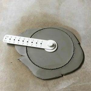 May include: A grey circular clay slab with a white plastic pottery tool centred on top. The tool features a rotating arm with numbered holes from 2 to 10, used for measuring and cutting precise circles in clay, resting flat on the surface.