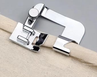 May include: A shiny, chrome-plated sewing machine foot, designed for hemming fabric. The foot is attached to a beige fabric, showing its function. The foot has a curved design with a screw for adjustment, and a flat base.