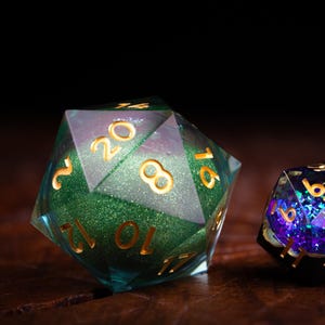 May include: Two polyhedral dice for tabletop games. One is a large, green and clear D20 with gold numbers. The other is a smaller, black D10 with purple and blue glitter. Both dice are on a wooden surface.