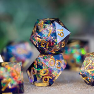 May include: A set of polyhedral dice with a shimmering, iridescent effect. The dice are primarily dark blue with gold numbering. The set includes a variety of shapes, including a 20-sided die, a 12-sided die, and a 4-sided die.