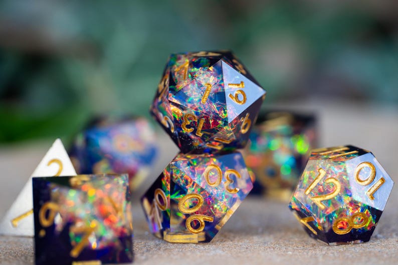 May include: A set of polyhedral dice with a shimmering, iridescent effect. The dice are primarily dark blue with gold numbering. The set includes a variety of shapes, including a 20-sided die, a 12-sided die, and a 4-sided die.