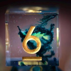 May include: A clear resin die with a gold number six and a gold bar. The interior features a swirling pattern of blue and green. The die is a unique and decorative item.