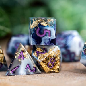 May include: A set of six polyhedral dice with a blue, white, and gold marbled design. The dice have purple numbers and are made of resin. The dice are on a wooden surface.