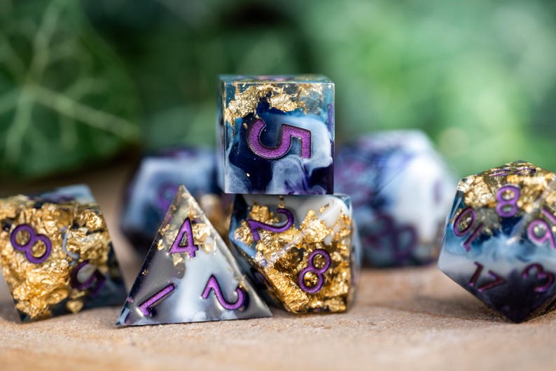 May include: A set of six polyhedral dice with a blue, white, and gold marbled design. The dice have purple numbers and are made of resin. The dice are on a wooden surface.