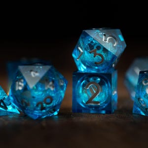 Op de afbeelding: Een verzameling doorschijnende blauwe dobbelstenen met zilveren cijfers. De dobbelstenen hebben verschillende vormen, waaronder een piramide, een kubus en veelzijdige vormen. De dobbelstenen hebben een wervelend, galaxy-achtig interieur.